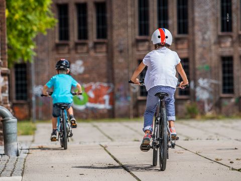Zwei Kinder fahren mit dem Fahrrad auf einem Weg vor einem Backsteingebäude mit Graffiti. Das Kind links trägt ein blaues Hemd und einen dunklen Helm, während das Kind rechts ein weißes Hemd und einen Helm mit einem roten Streifen trägt.