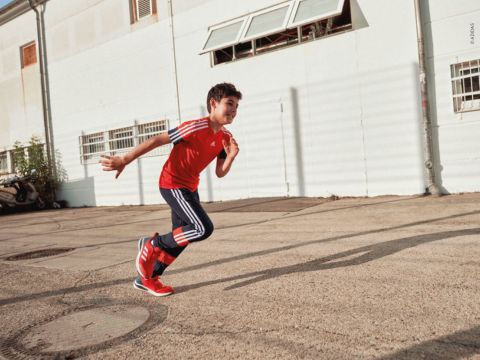 Ein kleiner Junge in rotem Hemd und blauer Hose läuft auf einer gepflasterten Fläche im Freien. Er wirkt voller Energie und steht in der Nähe eines hellen Gebäudes mit mehreren Fenstern. Auf dem Boden sind Schatten zu sehen, im Hintergrund sind ein kleiner Baum und ein geparkter Roller zu sehen.
