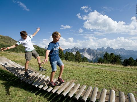 Zwei Jungen balancieren auf einer Holzbrücke in einer Graslandschaft mit Bergen und blauem Himmel im Hintergrund. Einer trägt ein weißes Hemd und Shorts, der andere ein blaues Hemd und Shorts. Sie wirken fröhlich und abenteuerlustig.