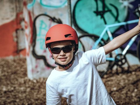 Ein junger Mensch mit rotem Helm und Sonnenbrille lächelt und streckt den Arm aus. Er steht vor Graffiti-bedeckten Wänden und einem Fahrrad und posiert auf spielerische Weise.