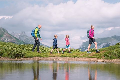 Eine vierköpfige Familie wandert auf einem malerischen, grasbewachsenen Pfad mit einem ruhigen Teich im Vordergrund. Im Hintergrund sind Berge unter einem teilweise bewölkten Himmel zu sehen. Jedes Mitglied trägt farbenfrohe Wanderkleidung und einen Rucksack.