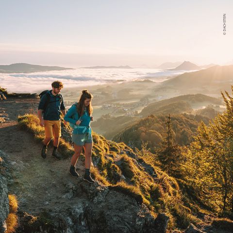Zwei Wanderer mit Rucksäcken gehen bei Sonnenaufgang einen grasbewachsenen Bergpfad entlang. Die Landschaft unter ihnen ist voller sanfter Hügel und eines Dorfes, das teilweise von Nebel bedeckt ist. Das Sonnenlicht taucht die Szene in ein warmes Licht.