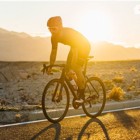 Ein Radfahrer fährt bei Sonnenuntergang auf einer asphaltierten Straße durch eine Berglandschaft. Die Sonne erzeugt hinter dem Fahrer, der Helm und Sonnenbrille trägt, einen warmen, leuchtenden Effekt. Die Landschaft ist in goldene Farbtöne getaucht.