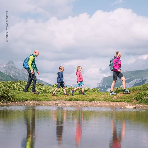 Eine vierköpfige Familie wandert auf einem Pfad in der Nähe eines Sees mit Spiegelungen im Wasser. Die Gruppe besteht aus zwei Erwachsenen und zwei Kindern, alle tragen Rucksäcke. Sie sind von üppigen grünen Hügeln umgeben und im Hintergrund ist ein bewölkter Himmel.