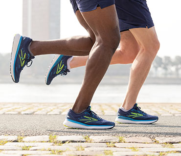 Zwei Personen laufen auf einem gepflasterten Weg in der Nähe eines Gewässers. Sie tragen blaue Sportschuhe mit Neonakzenten und dunkle Shorts. Der Fokus liegt auf ihren Beinen und Füßen.