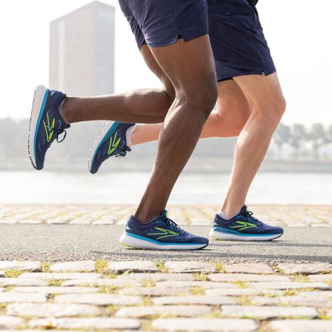 Zwei Personen joggen auf einem gepflasterten Weg in der Nähe eines Gewässers. Beide tragen blaue Laufschuhe und Shorts. Im Hintergrund ist ein hohes Gebäude zu sehen, und Bäume säumen den Horizont. Der Fokus liegt auf ihren Beinen und Schuhen.