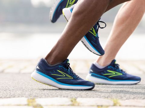 Nahaufnahme von zwei Personen, die auf einem gepflasterten Weg joggen. Sie tragen blaue Laufschuhe mit grünen und weißen Akzenten. Der Hintergrund ist verschwommen, was Bewegung suggeriert.