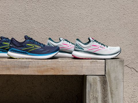 Drei Paar Sportschuhe stehen auf einer Holzbank vor einer strukturierten beigen Wand. Die Schuhe sind marineblau mit gelben Akzenten, blaugrün mit rosa Akzenten und hellgrau mit rosa Akzenten und haben alle gepolsterte Sohlen.