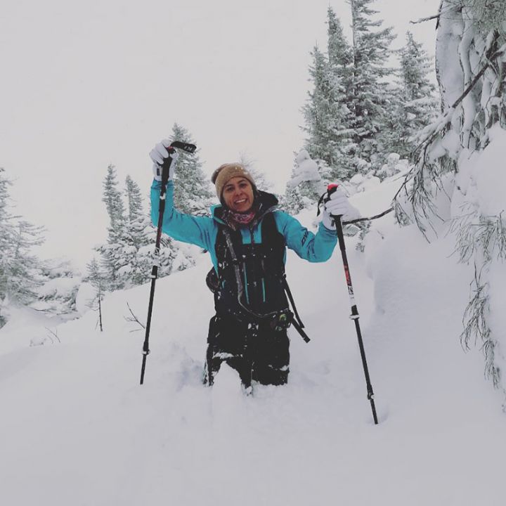 Eine lächelnde Person steht im tiefen Schnee und hält Skistöcke in der Hand. Sie trägt Winterkleidung, darunter eine blaue Jacke und eine beige Mütze. Um sie herum sind schneebedeckte Bäume und eine Berglandschaft. Der Himmel ist bedeckt.
