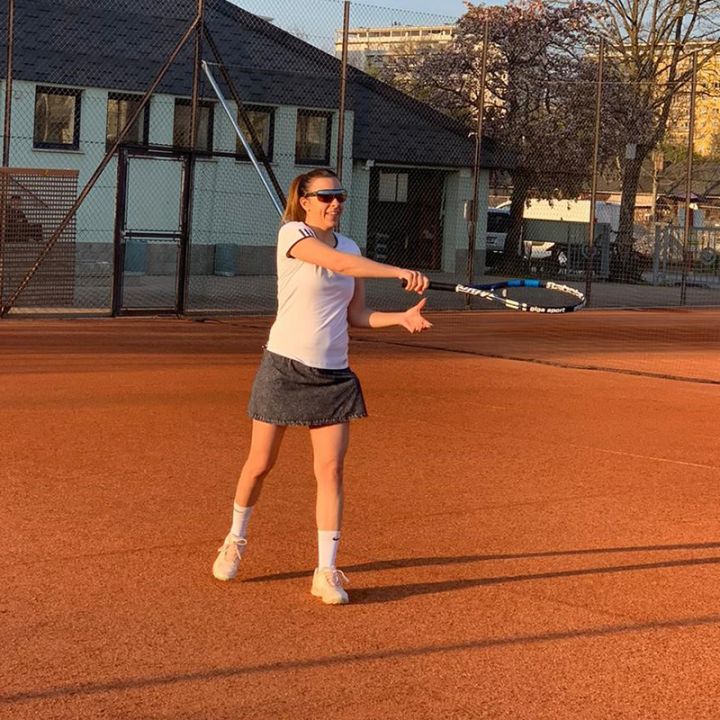 Eine Person spielt bei Sonnenuntergang Tennis auf einem Sandplatz. Sie trägt ein weißes Hemd, einen dunklen Rock und eine Sonnenbrille. Im Hintergrund sind ein Gebäude und ein eingezäunter Bereich zu sehen.