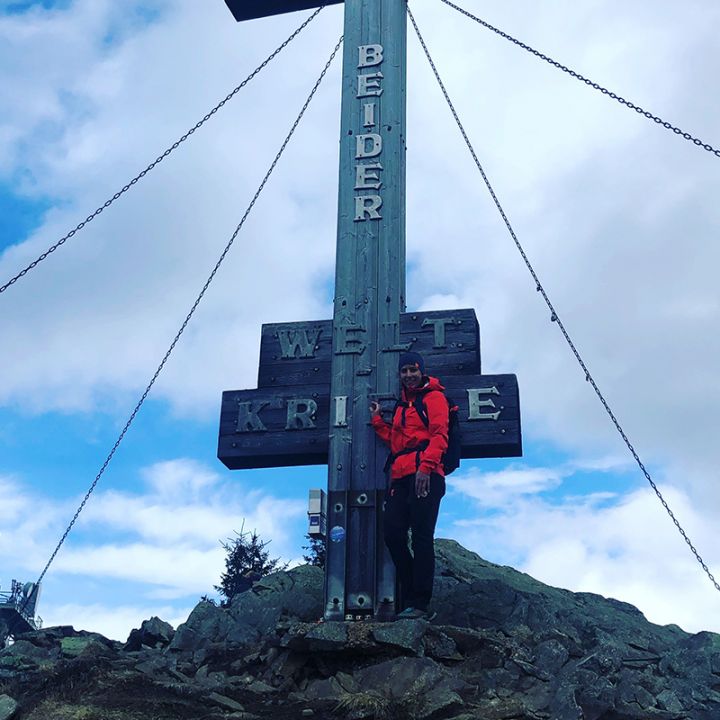 Eine Person in einer roten Jacke steht auf dem Gipfel eines Berges und lehnt an einem großen Holzkreuz mit Metallketten. Der Himmel ist bewölkt und im Hintergrund ist ein kleines Gebäude zu sehen.