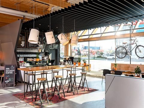 Modernes Café-Interieur mit langem Stehtisch und Hockern. Hängelampen über dem Tisch, ein an der Wand befestigtes Fahrrad und Blick auf die Straße durch große Fenster. Auf der linken Seite befinden sich eine Kaffeestation und ein Schild, überall farbenfrohes Dekor.