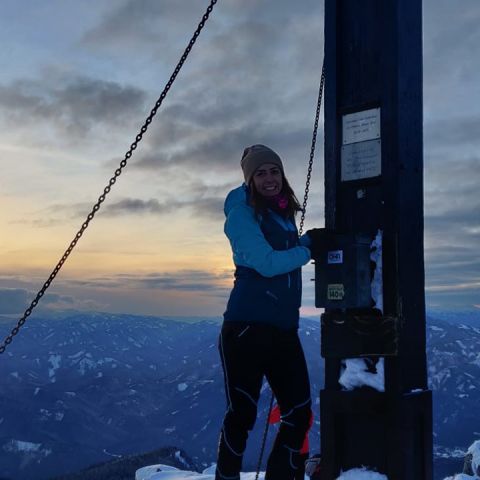 Eine Person mit einer blauen Jacke und einer beigen Mütze steht bei Sonnenuntergang neben einem Holzpfosten auf einem schneebedeckten Berggipfel. Der Himmel ist in einer Mischung aus Orange- und Blautönen gehalten, im Hintergrund sind Bergketten zu sehen.