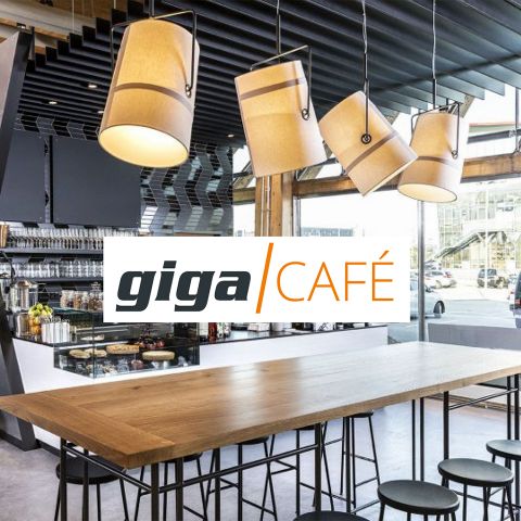 Innen ein modernes Café mit einem langen Holztisch und hohen Hockern. Große, zylindrische Pendelleuchten hängen darüber. Das Café hat eine Glasfront und auf einem Schild steht in fettem, orangefarbenem Text „Giga Café“.