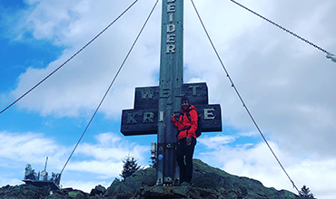 Eine Person in einer roten Jacke steht neben einem großen Metallkreuz auf einem Berggipfel. Auf dem Kreuz steht die Aufschrift „GLETSCHERKREUZ“. Der Himmel ist teilweise bewölkt und im Hintergrund sind weitere Berggipfel zu sehen.