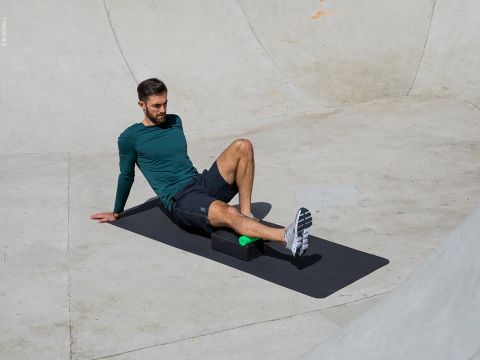 Eine Person in einem langärmeligen grünen Hemd und schwarzen Shorts sitzt auf einer schwarzen Matte und massiert ihre Wade mit einem Schaumstoffblock und einem grünen Ball. Die Szene scheint eine Betonfläche zu sein, möglicherweise ein Skatepark oder eine Sportanlage.