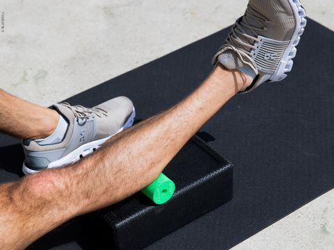Eine Person dehnt ihre Wade auf einer schwarzen Schaumstoffrolle, trägt graue Sportschuhe und sitzt auf einer Trainingsmatte. Die Umgebung ist eine neutrale, helle Fläche.