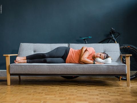 Eine Person in Sportkleidung schläft auf einem grauen Sofa mit einem weißen Kissen, die Arme verschränkt, im Hintergrund ein Fahrrad und darunter ein Parkettboden.