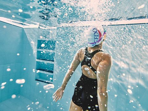 Ein Schwimmer in einem schwarzen Badeanzug mit weißer Badekappe liegt unter Wasser in einem Pool. Er ist von Blasen umgeben und blickt nach oben, während Sonnenlicht durch das Wasser fällt. Im Hintergrund ist eine geflieste Wand zu sehen.