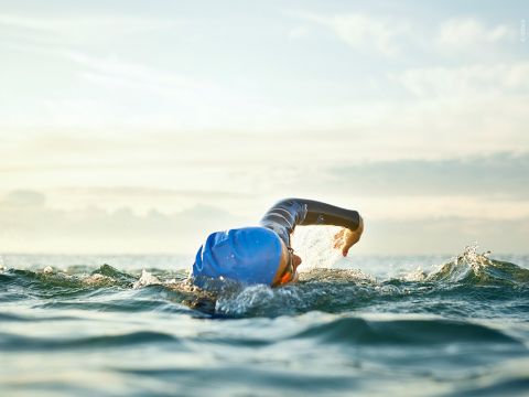 Ein Schwimmer mit blauer Badekappe und Schwimmbrille schwimmt im offenen Wasser Kraulstil. Die sonnenbeschienenen Wellen plätschern um sie herum und der Horizont ist unter einem sanften, bewölkten Himmel sichtbar.