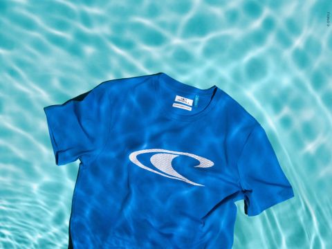 Ein blaues T-Shirt mit einem weißen Wellenlogo, das auf der Oberfläche eines klaren, türkisfarbenen Pools schwimmt. Das Licht erzeugt schimmernde Muster auf dem Wasser.