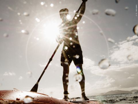 Eine Person fährt Stand-Up-Paddleboard auf dem Meer. Die Silhouette ist vor einer hellen Sonne zu sehen, im Vordergrund sind Wassertropfen zu sehen. Der Himmel ist teilweise bewölkt und am Horizont sind in der Ferne Berge zu sehen.