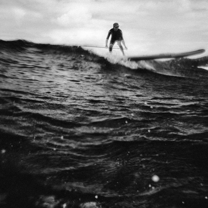 Schwarzweißbild einer Person, die auf einer großen Meereswelle paddelt. Die Perspektive ist niedrig und fängt die Textur des Wassers im Vordergrund ein. Die Person steht und wirkt konzentriert, im Hintergrund ist hügeliges Land zu sehen.