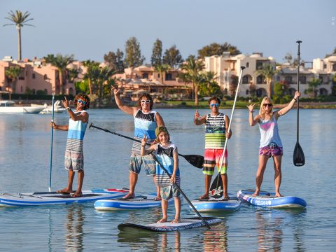 Eine Gruppe von fünf Personen in farbenfroher Strandkleidung und Sonnenbrillen steht auf Paddleboards in einem ruhigen Gewässer. Sie lächeln und gestikulieren mit den Händen. Im Hintergrund sind Palmen und Gebäude zu sehen.