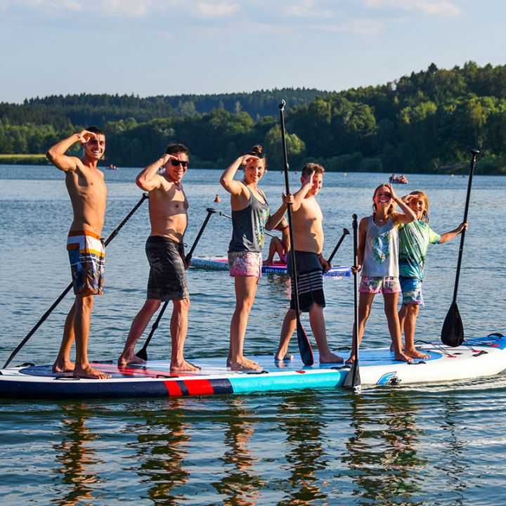 Sechs Personen stehen auf einem großen Paddleboard auf einem See, jeder hält ein Paddel und lächelt. Sie tragen Badeanzüge und sind von ruhigem Wasser und einer malerischen Aussicht auf Bäume und einen klaren Himmel umgeben.