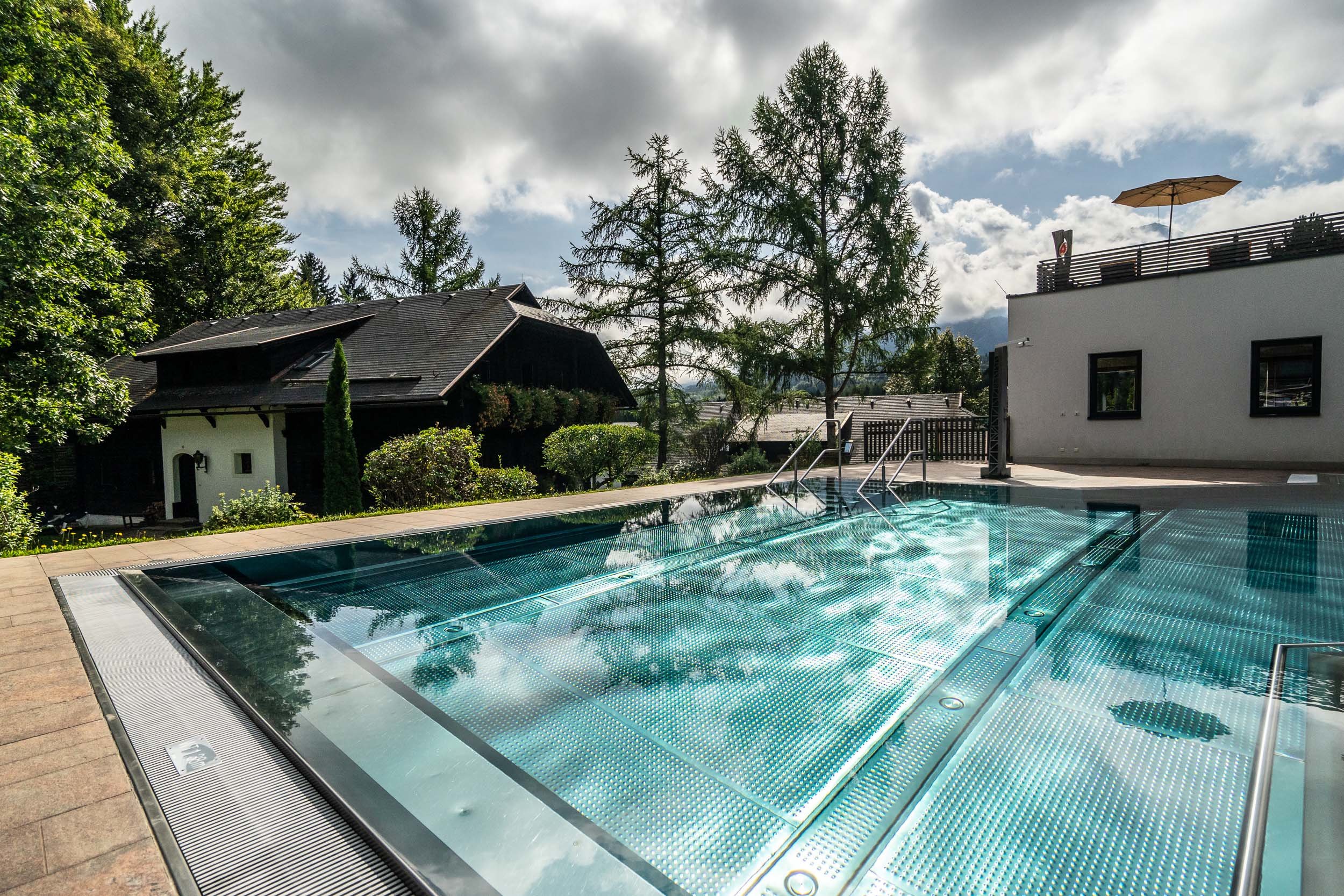 Ein moderner Außenpool mit transparentem Boden, der den wolkigen Himmel reflektiert. Um ihn herum sind hohe Bäume und ein Holzhaus mit schrägem Dach. Im Hintergrund ist ein weißes Gebäude mit einem Sonnenschirm auf dem Dach zu sehen.