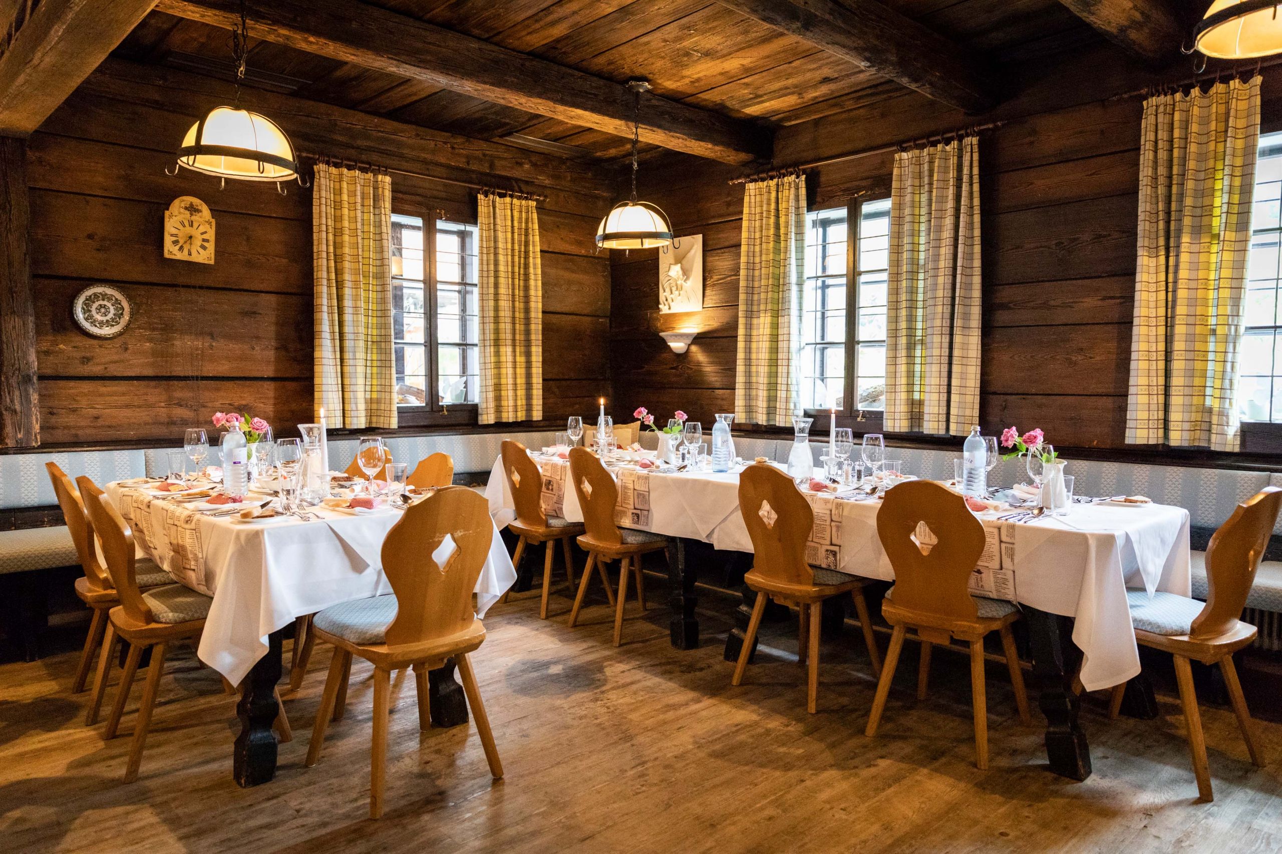 Ein gemütliches Esszimmer aus Holz mit zwei Tischen, die für eine Mahlzeit gedeckt sind. Die Tische sind mit weißen Tischdecken, Glaswaren und Blumenschmuck geschmückt. Die Tische sind von Holzstühlen umgeben. Die Fenster haben karierte Vorhänge und der Raum verfügt über eine warme, rustikale Beleuchtung.