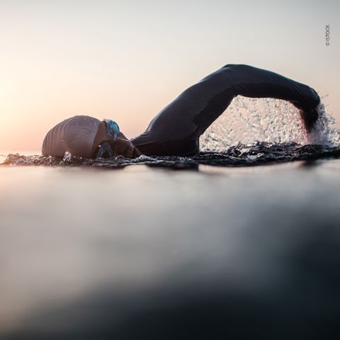 Ein Schwimmer in Neoprenanzug und Schwimmbrille schwimmt bei Sonnenaufgang im offenen Wasser Kraulstil. Der Arm ist über dem Wasser ausgestreckt, Tropfen spritzen herum und fangen die Bewegung und Energie ein.