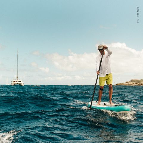 Eine Person, die auf dem Meer paddelt, trägt ein weißes Langarmshirt, gelbe Shorts und einen Hut. Das Meer ist leicht wellig und im Hintergrund ist unter einem teilweise bewölkten Himmel ein Segelboot zu sehen.
