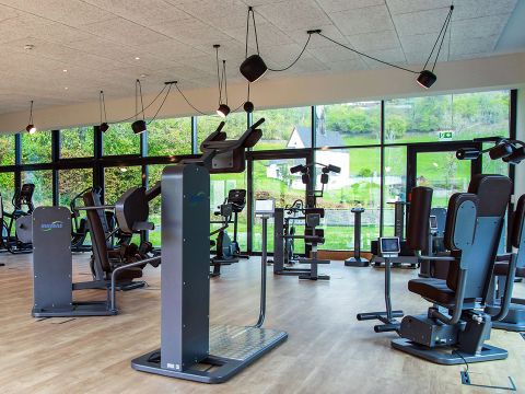 Ein modernes Fitnessstudio mit großen Fenstern, die einen malerischen Blick nach draußen bieten. Das Fitnessstudio ist mit verschiedenen Fitnessgeräten ausgestattet, darunter Laufbänder, Crosstrainer und Kraftgeräte. Der Boden ist aus Holz und darüber hängen Deckenleuchten.