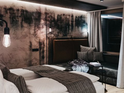 Modernes Schlafzimmer mit großem Bett, braun gepolstertem Kopfteil und künstlerischem schwarz-weißem Wandgemälde. Sanfte Beleuchtung, eine gemütliche Bank, Kissen und ein Beistelltisch mit Büchern tragen zur warmen Atmosphäre bei. Vorhänge rahmen ein dunkles Fenster ein.