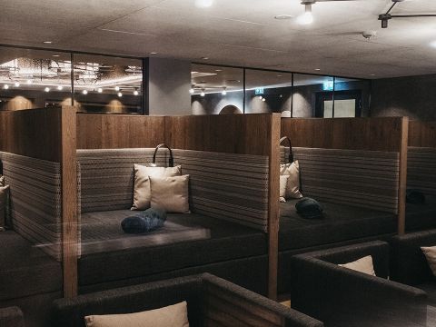 Ein moderner Loungebereich mit Holzkabinen, jede mit gestreiften Kissen, grauen gerollten Handtüchern und sanfter Umgebungsbeleuchtung. Die Kabinen sind durch Holztrennwände voneinander getrennt und der Raum ist auf Entspannung und Komfort ausgelegt.