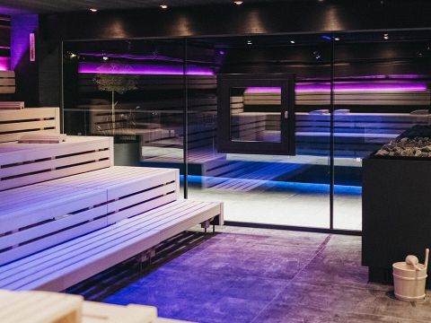 Moderne Saunaeinrichtung mit Holzbänken und warmer Beleuchtung. Eine Glaswand trennt die Sauna von einem anderen Bereich, und violette Akzentlichter sorgen für eine entspannende Atmosphäre. Ein Eimer und eine Schöpfkelle stehen rechts neben den Ecksteinen.