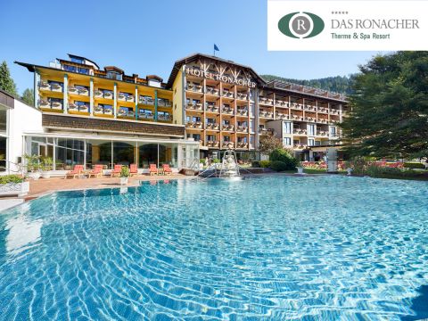 Ein luxuriöses Hotel mit einem großen Außenpool, der von Liegestühlen umgeben ist. Das Gebäude verfügt über Balkone und eine Glasfassade. Im Hintergrund sind Bäume und Berge zu sehen. Auf dem Schild steht „Hotel Ronacher“ und das Logo „Das Ronacher Therme & Spa Resort“.