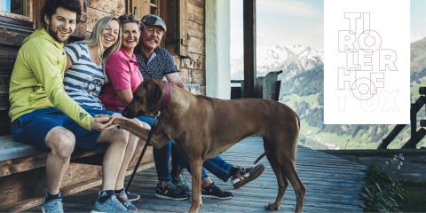 Eine Gruppe von vier Personen sitzt auf einer Holzbank vor einer Hütte, lächelt und interagiert mit einem großen braunen Hund. Im Hintergrund sind schneebedeckte Berge zu sehen. Der Text rechts lautet „TIROLERHOF TUX“.