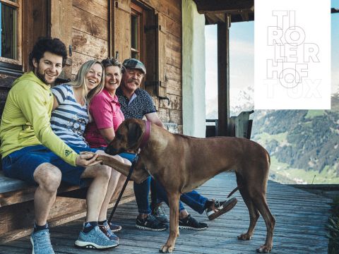 Eine Gruppe von vier Personen sitzt auf einer Holzveranda, vor ihnen ein großer Hund. Sie lächeln und blicken in die Kamera. Im Hintergrund sind Berge zu sehen und auf der rechten Seite des Bildes ein Logo.