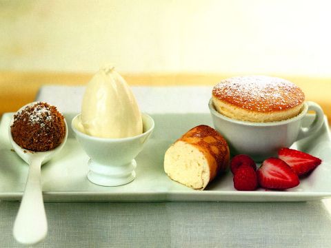 Eine Dessertplatte mit einem kleinen Soufflé in einer mit Puderzucker bestäubten Tasse, einer Kugel Vanilleeis in einer Schüssel, einer Pralinenkugel auf einem Löffel, einem gerollten Kuchenstück und frischen Himbeer- und Erdbeerscheiben auf einem rechteckigen Teller.