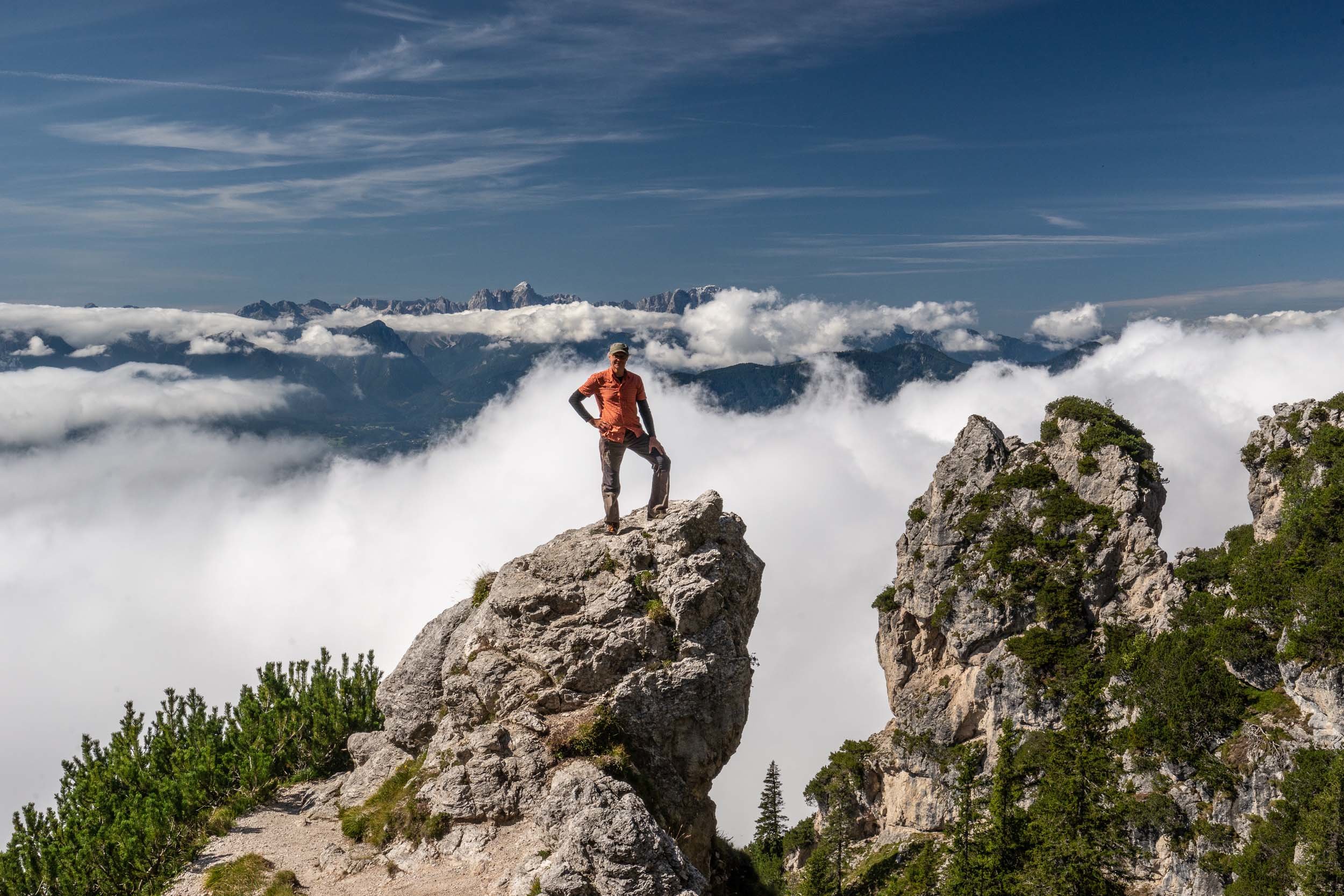 Eine Person steht selbstbewusst auf einem felsigen Gipfel, umgeben von Wolken und üppigem Grün, mit einer riesigen Bergkette im Hintergrund unter einem klaren blauen Himmel.