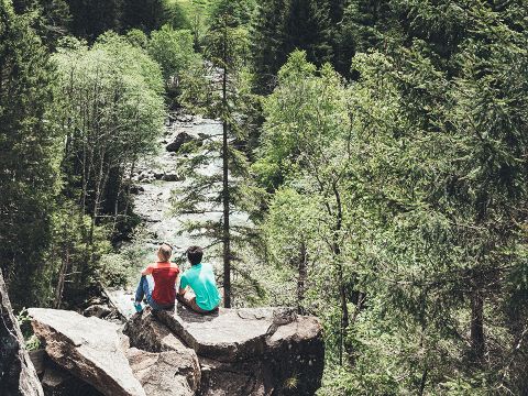 Zwei Menschen sitzen auf einem großen Felsen und blicken auf einen üppigen, grünen Wald, durch den ein Bach fließt. Hohe Bäume umgeben sie und schaffen eine friedliche und malerische Szene in der Wildnis.