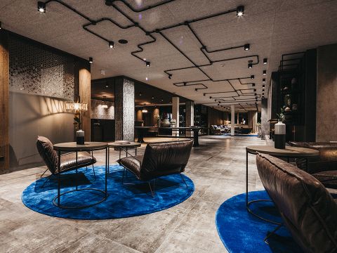 Moderne Hotellobby im Industriedesign mit Lederstühlen um runde Tische auf hellblauen Teppichen. Sichtbare Deckenrohre und sanfte Beleuchtung sorgen für eine gemütliche Atmosphäre. Im Hintergrund sind eine Rezeption und ein Loungebereich zu sehen.