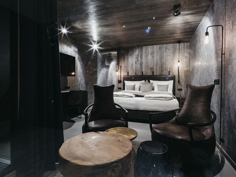 Ein modernes, schwach beleuchtetes Schlafzimmer mit rustikalem Holzthema. Es verfügt über ein großes Bett mit weißer Bettwäsche, zwei dunkle Lederstühle, einen runden Couchtisch aus Holz und einen an der Wand montierten Fernseher. Dezente Beleuchtung trägt zur gemütlichen Atmosphäre bei.