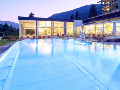Im Abendlicht leuchtender Außenpool, umgeben von einem modernen Gebäude mit großen Fenstern und einer Bergkulisse. Auf der angrenzenden Rasenfläche sind Liegestühle am Pool aufgereiht.