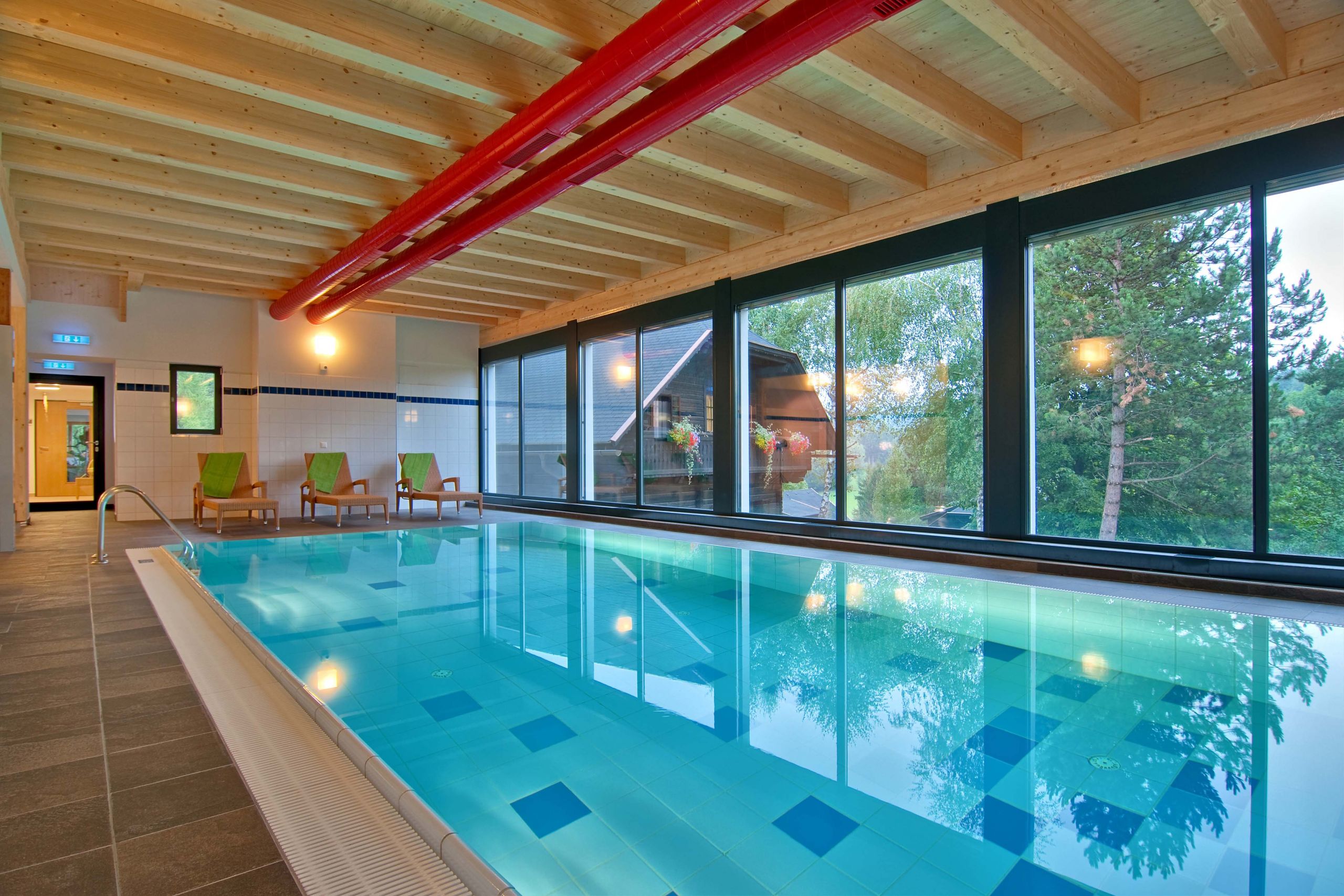 Innenpool mit blauem Wasser und Holzbalken an der Decke. Große Fenster auf einer Seite bieten einen Blick auf Bäume und Grün draußen. Liegestühle mit grünen Kissen sind an der Seite angeordnet und Deckenleuchten beleuchten den Bereich.