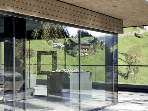 Modernes Gebäude mit großen Glasfenstern, die einen malerischen Blick auf grasbewachsene Hügel, Bäume und ein rustikales Holzhaus bieten. Die Inneneinrichtung umfasst minimalistische Möbel und einen Blumentopf. Die Holzdecke bildet einen Kontrast zum glatten Glas.