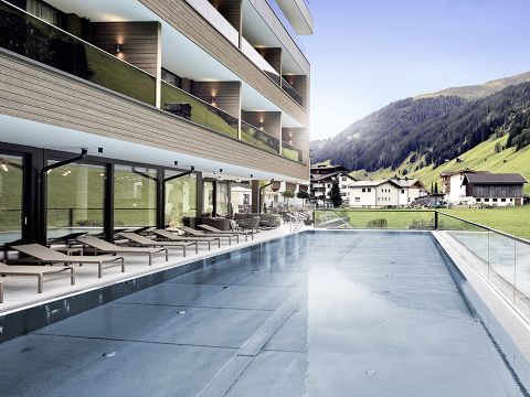 Ein modernes Hotel mit elegantem Design verfügt über einen Außenpool, der von Liegestühlen umgeben ist. In den Glasfenstern des Gebäudes spiegeln sich der Pool und die nahe gelegene Rasenfläche wider, während sich im Hintergrund Berge unter einem klaren blauen Himmel erheben.