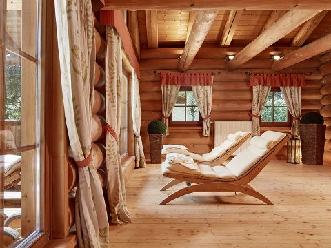Ein gemütlicher Raum mit Wänden und Decke aus Holzbalken verfügt über zwei gepolsterte Loungesessel mit Handtüchern auf einem Holzboden. Große Fenster mit Vorhängen lassen natürliches Licht herein und schaffen eine warme und einladende Atmosphäre. Topfpflanzen schmücken den Raum.